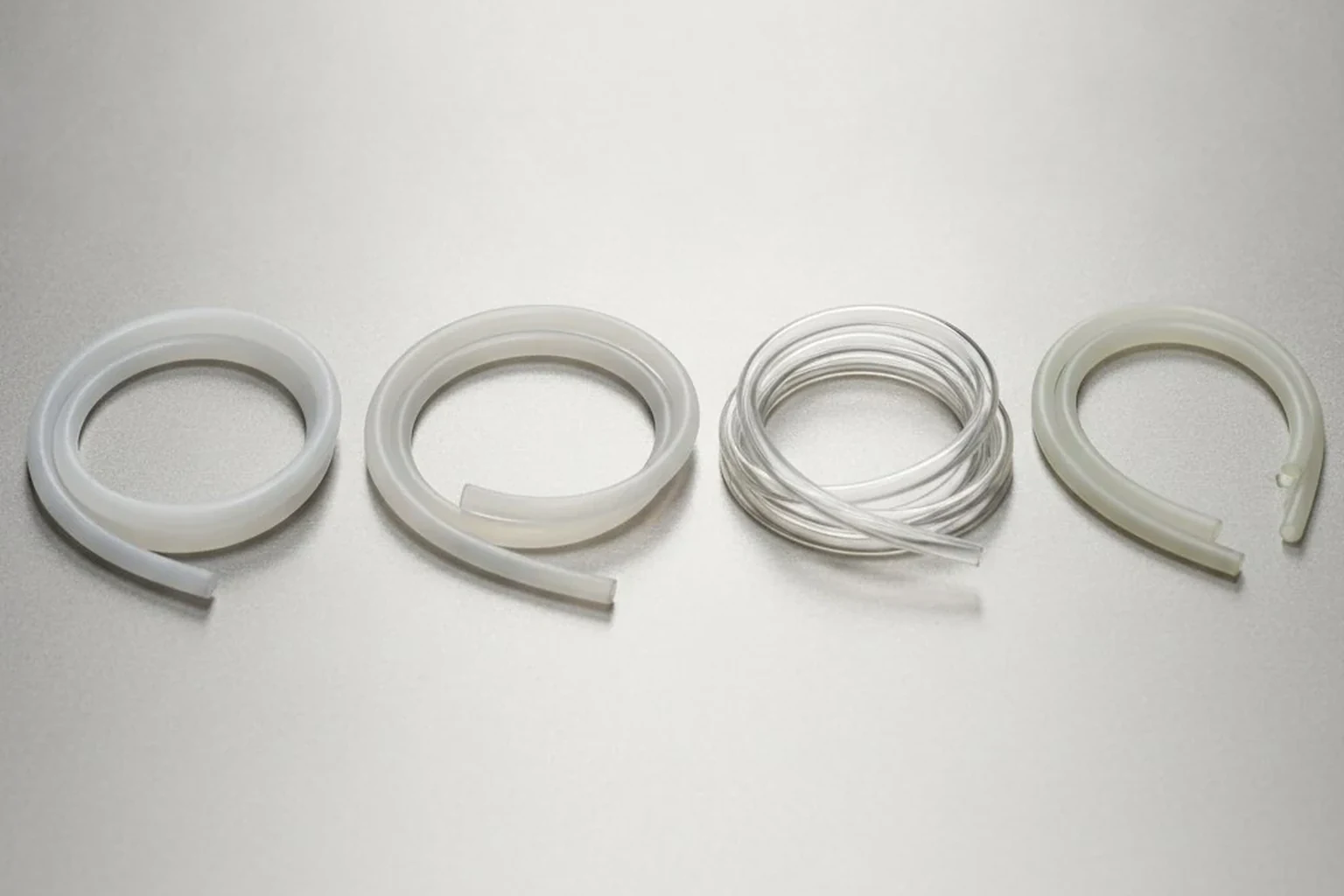 Photorealistic B2B industrial website image comparing PTFE, PFA, FEP and ETFE tubing by appearance and flexibility, four tubing samples arranged on a clean surface